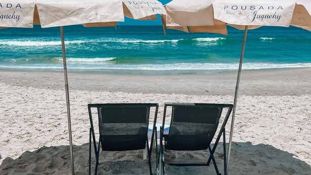 Two beach umbrellas with "Pousada" branding, a metal chair, and a sandy beach with ocean waves in the background. - Hiu Hotel (São Sebastião)