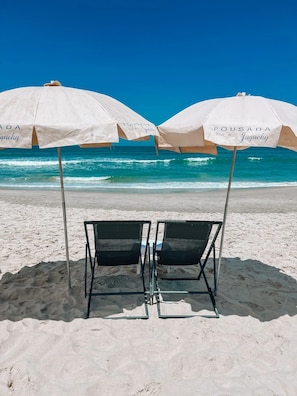 Two beach umbrellas with "Pousada" branding, a metal chair, and a sandy beach with ocean waves in the background. - Hiu Hotel (São Sebastião)