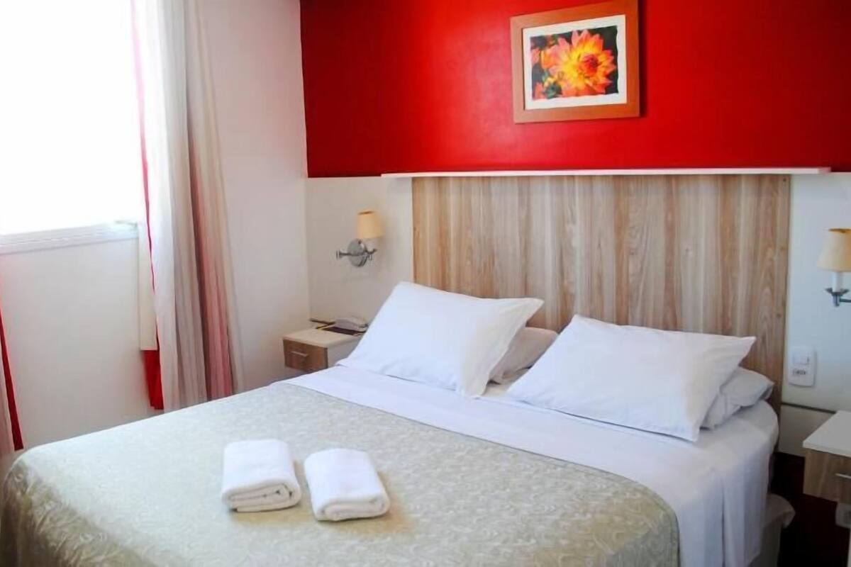 A hotel room with a bed, two pillows, a nightstand, a lamp, and a window with curtains.