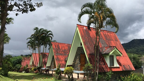A row of triangular-roofed buildings with a stone base, surrounded by lush greenery and palm trees. - Phunaya Resort (Khao Kho)
