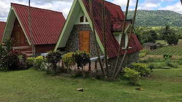 A bungalow with a stone foundation and a red metal roof, surrounded by lush greenery and a hilly landscape.