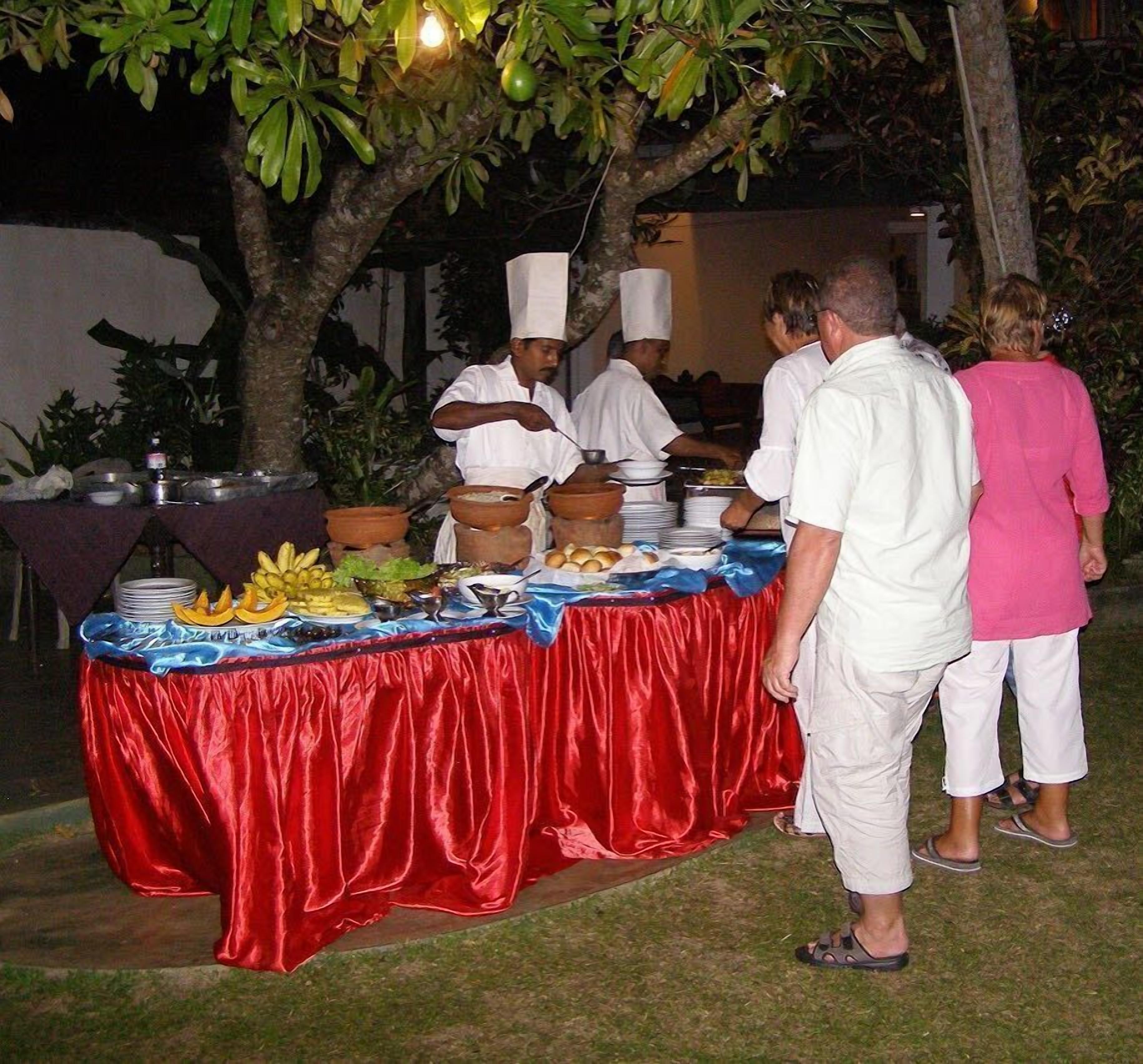 Disposição de buffet com toalha vermelha, diversos alimentos e pessoas servindo.