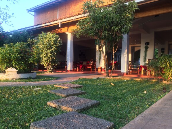 A covered outdoor seating area with stone steps leading to a patio, surrounded by greenery and a building with a tiled roof.