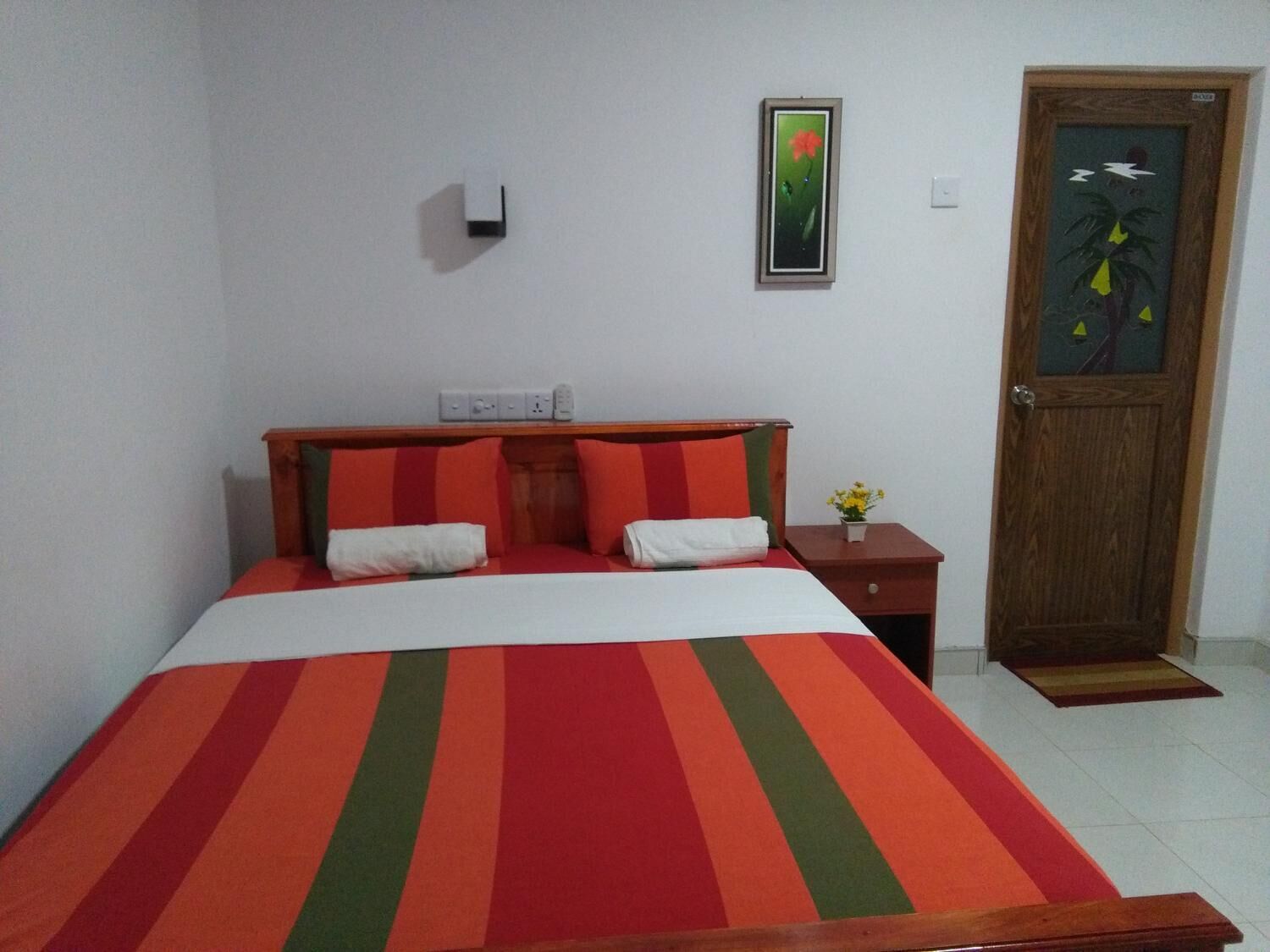 A bed with a striped bedspread, a wooden headboard, and two pillows. There is a nightstand with a vase of flowers and a framed picture on the wall.