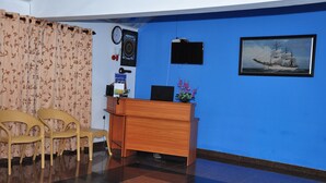 A reception area with a blue wall, a wooden desk, two wicker chairs, a clock, a framed picture of a ship, and a potted plant. - Hotel Camorich (Thissamaharama)