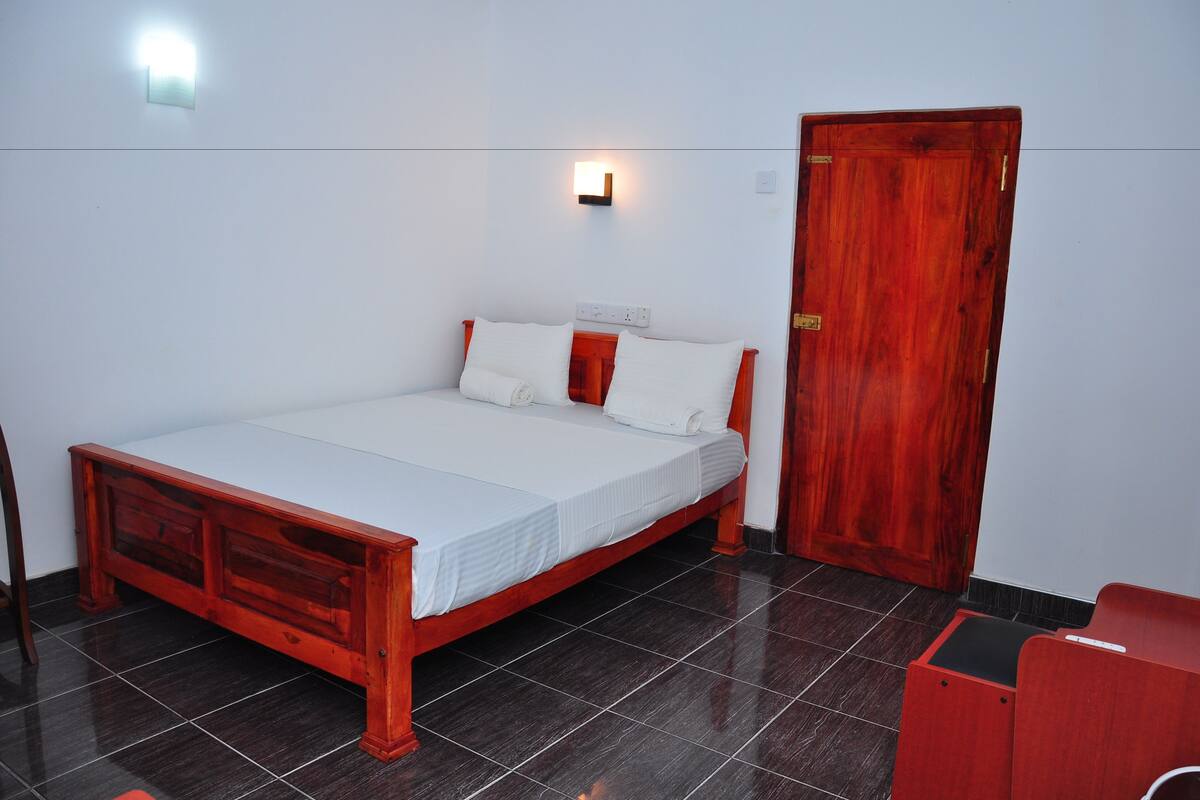 A single-bed room with a wooden frame, white bedding, a red bench, a wooden door, and a wall-mounted light fixture.
