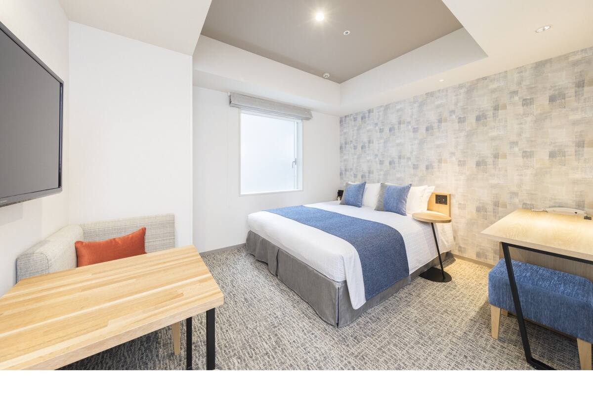 Ein modernes Hotelzimmer mit einem Bett, einem Schreibtisch, einem Stuhl, einem Fenster und einer Wand mit strukturierter Oberfläche.