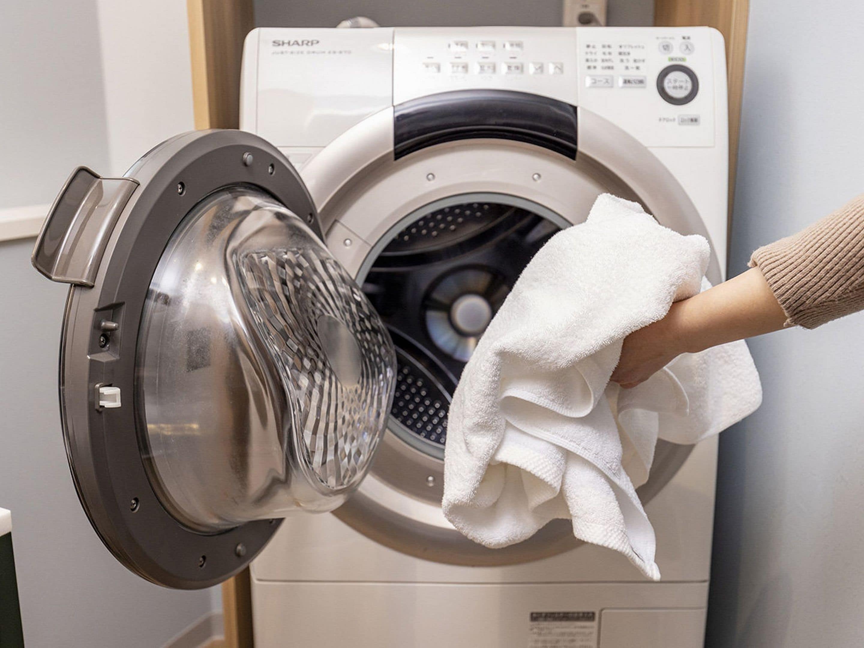A front-loading washing machine with a transparent door, a control panel with various buttons, and a white towel hanging on the door.
