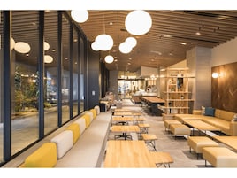 Un café moderno con mesas de madera, asientos con cojines amarillos, ventanas amplias e iluminación colgante.