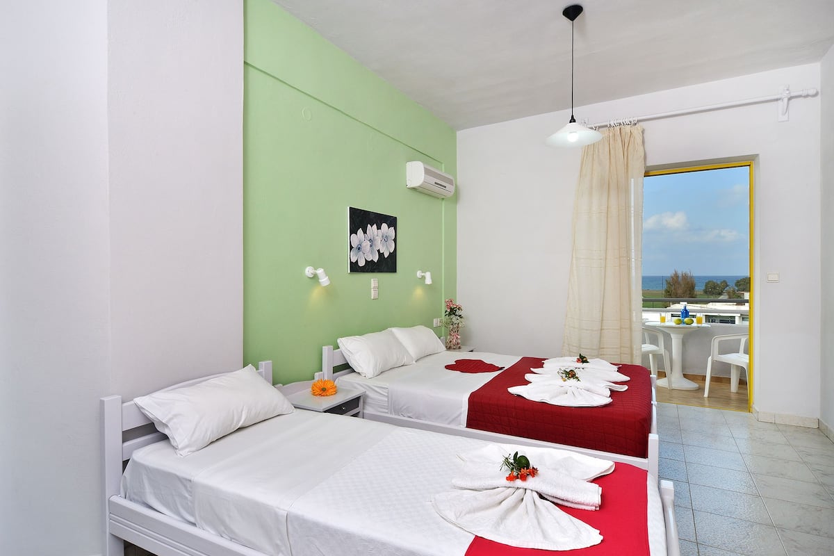 A hotel room with two beds, a green accent wall, an air conditioning unit, a window with a view, and a table with chairs outside.