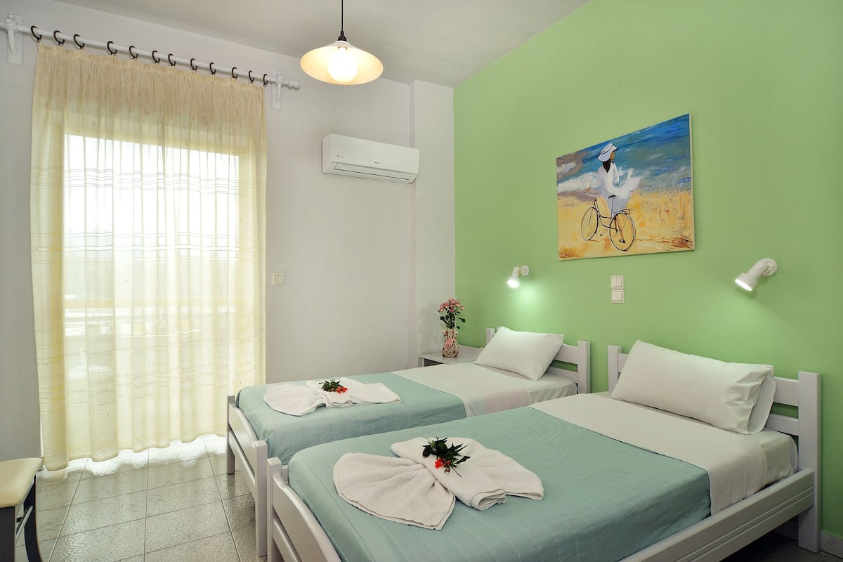 A hotel room with two beds, a window with curtains, an air conditioner, a painting on the wall, and a bedside lamp.