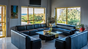 A modern living room with a sectional sofa, a coffee table, and large windows offering a view of the outdoors. - Nereides Hotel (Platanias)