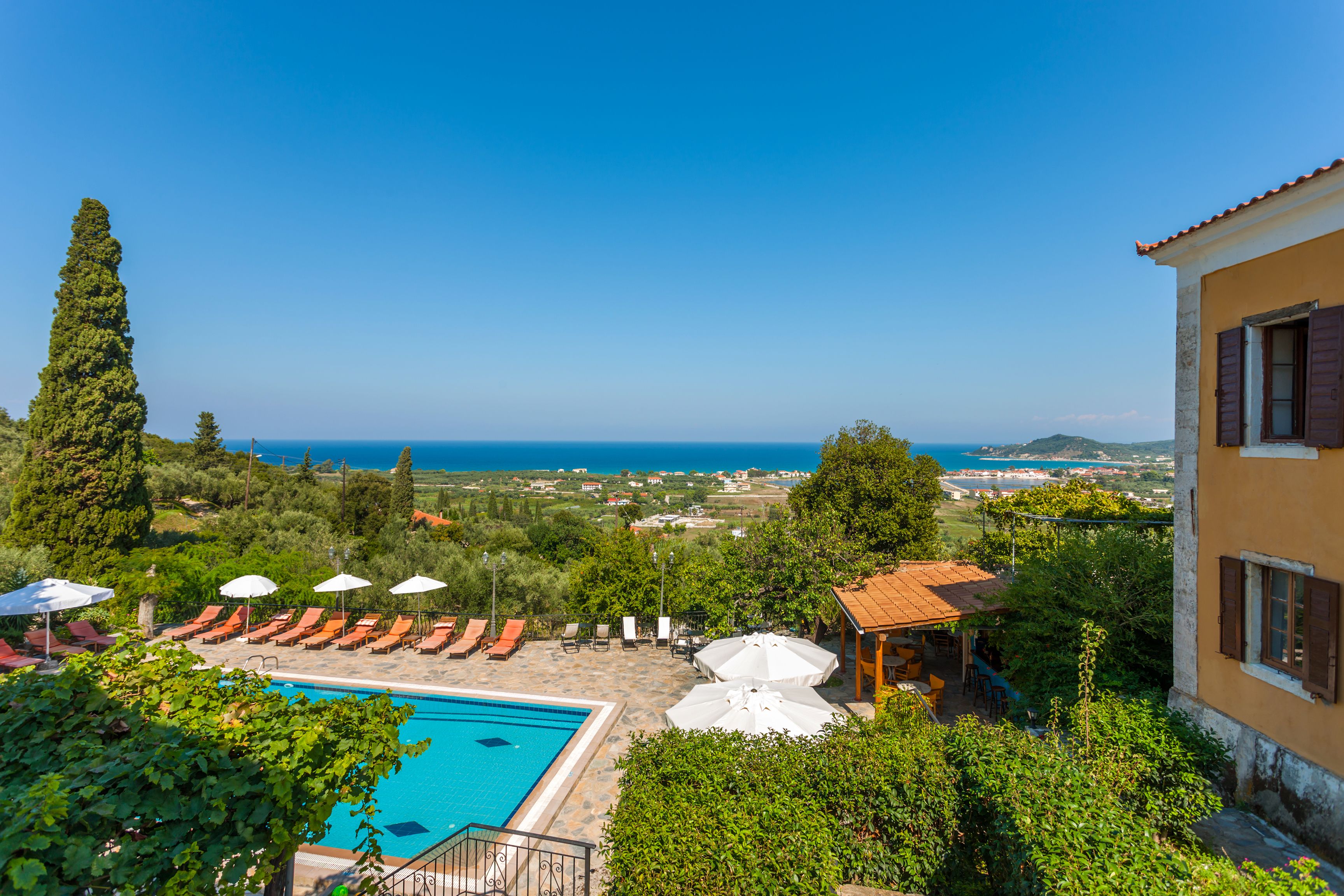 A pool with lounge chairs and umbrellas, a view of the sea, and a building with a tiled roof.