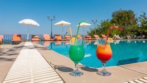 Dos cócteles sobre una mesa junto a una piscina, con sombrillas y sillones reclinables al fondo.