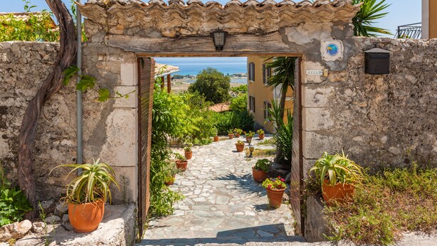 Un sendero de piedra conduce a una construcción rústica con tejado de tejas, rodeada de macetas con plantas y vistas al mar.