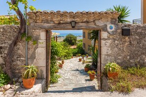 Un sendero de piedra conduce a una construcción rústica con tejado de tejas, rodeada de macetas con plantas y vistas al mar.