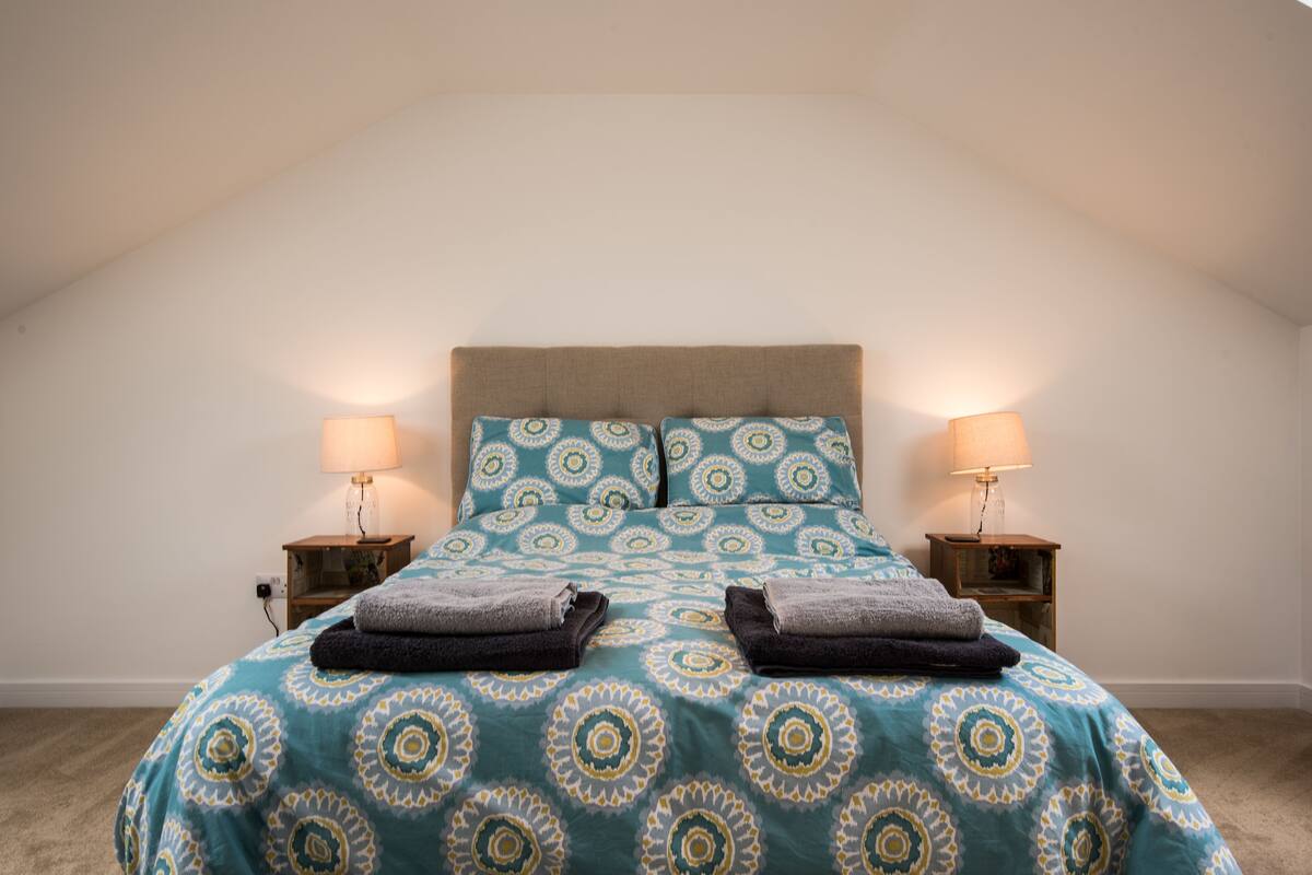 Une chambre à coucher avec une couvre-lit à motifs, deux oreillers, deux tables de chevet avec des lampes et des serviettes pliées sur le lit.