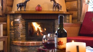 A cozy cabin interior with a stone fireplace, wooden walls, and a dining table set with a bottle, glasses, and fruits.