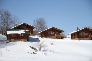 A row of log cabins with snow-covered roofs and stone foundations, set against a clear blue sky.