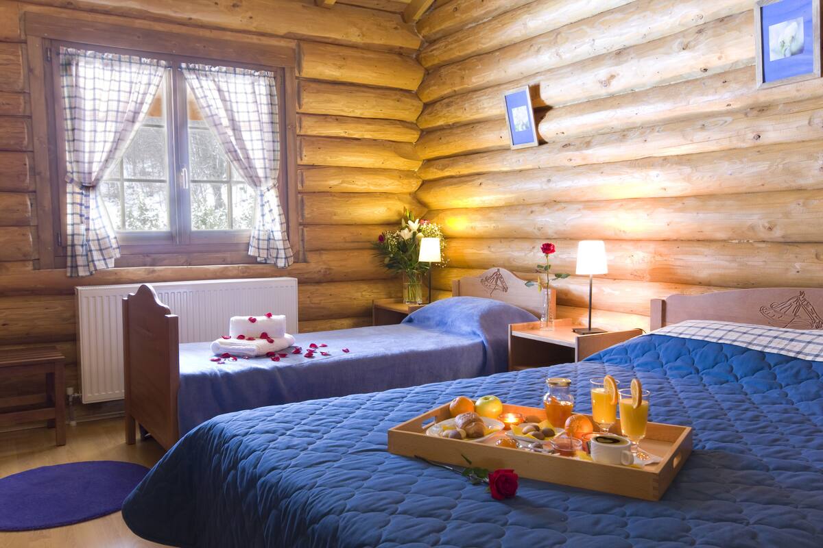Una habitación de cabaña de madera con dos camas, piso de madera, una ventana con cortinas, una mesita de noche con lámpara y una bandeja con alimentos para el desayuno.