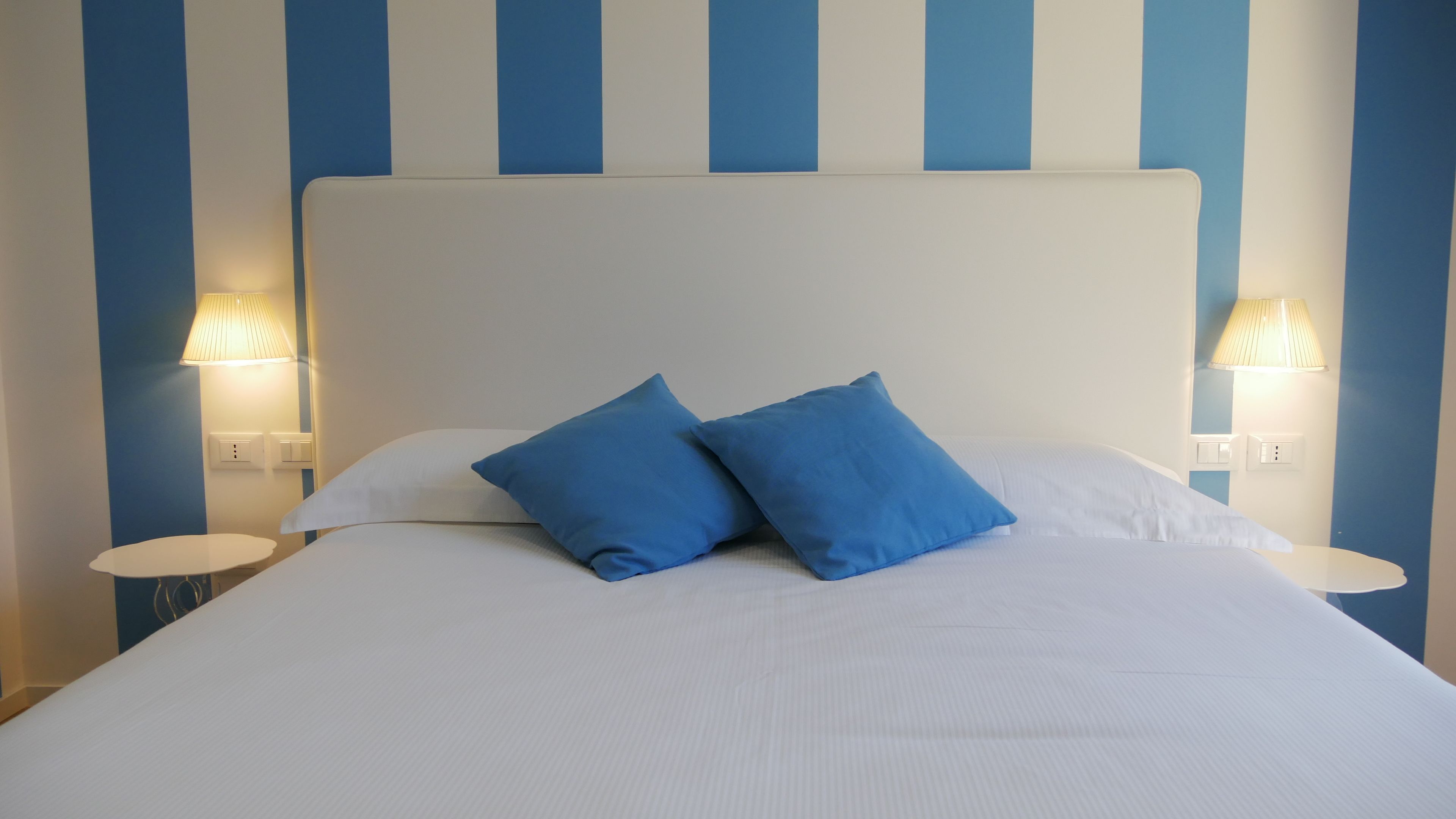A bed with white bedding and two blue pillows, flanked by two bedside lamps with white bases and beige shades.