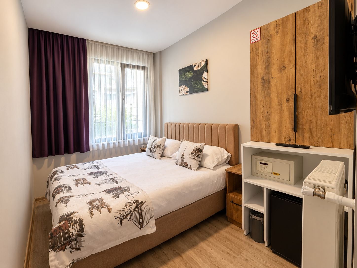 A hotel room with a bed, a nightstand, a wooden wardrobe, a television, and a small shelf with a trash bin.