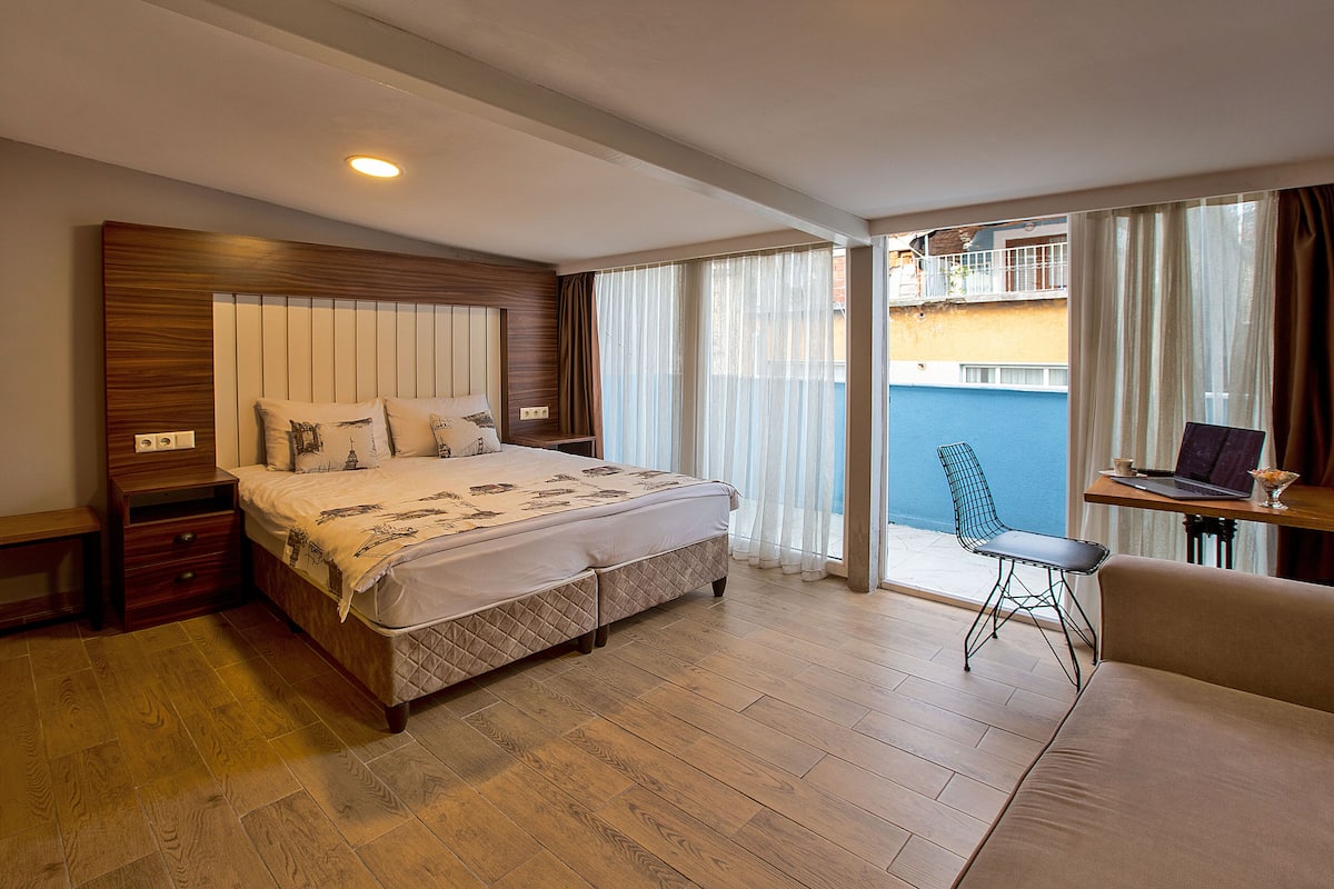 A hotel room with a large bed, a desk with a laptop, a chair, and a balcony with curtains.