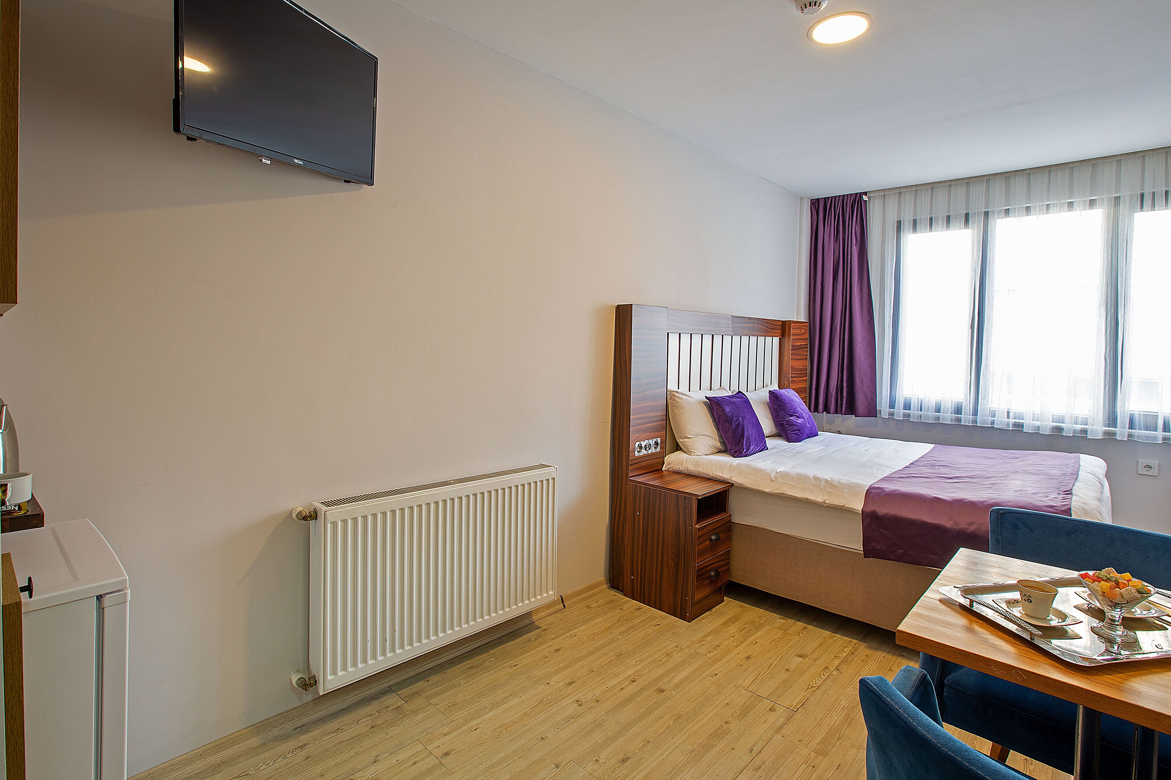 A hotel room with a bed, a TV mounted on the wall, a window with curtains, a radiator, a table with a tray, and a chair.