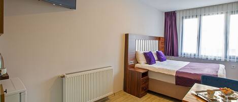 A hotel room with a bed, a TV mounted on the wall, a window with curtains, a radiator, a table with a tray, and a chair.