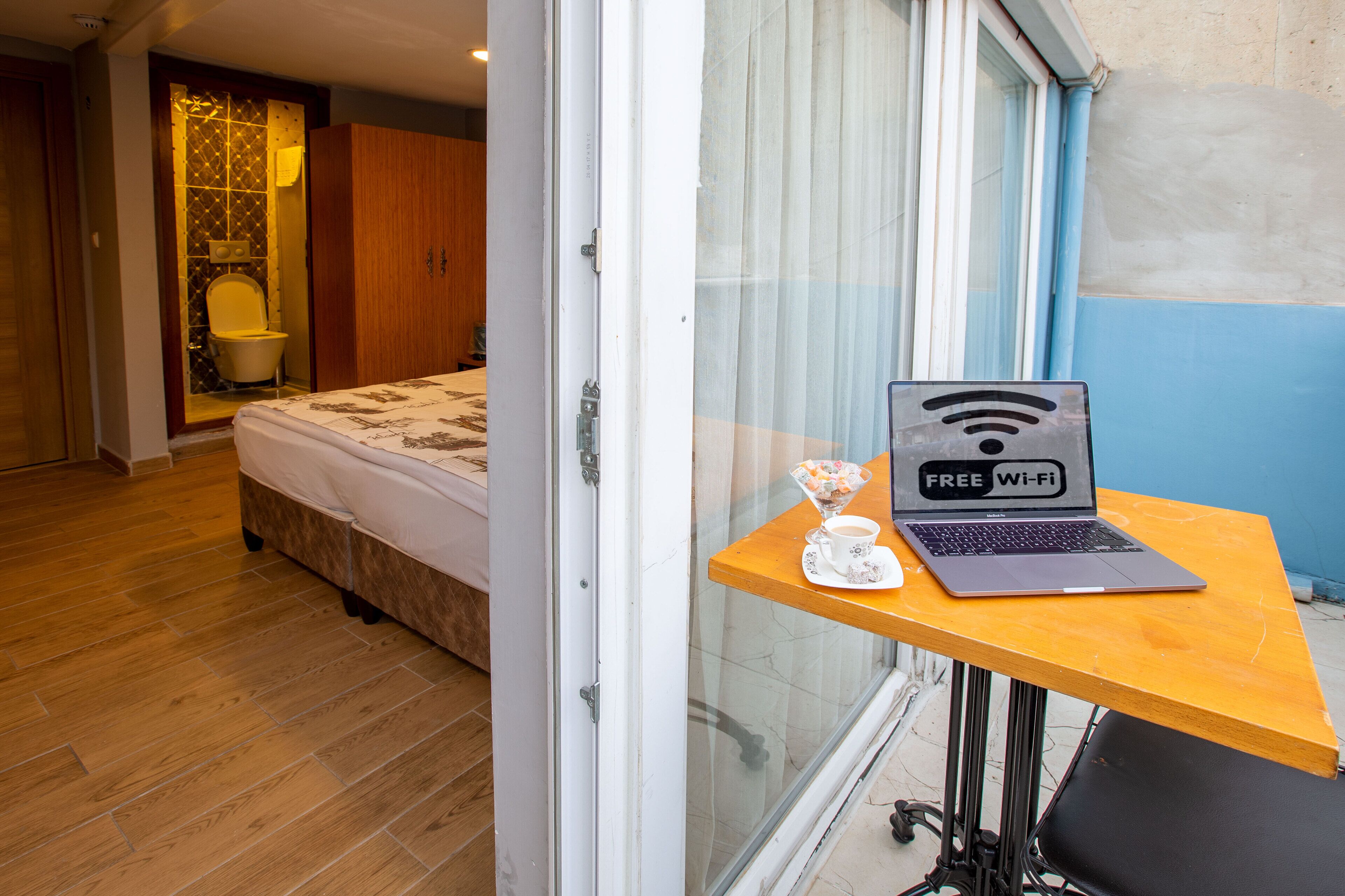 Une chambre d’hôtel avec un lit, un plancher en bois, une salle de bain avec des toilettes et un lavabo, ainsi qu’une table avec un ordinateur portable et une tasse.