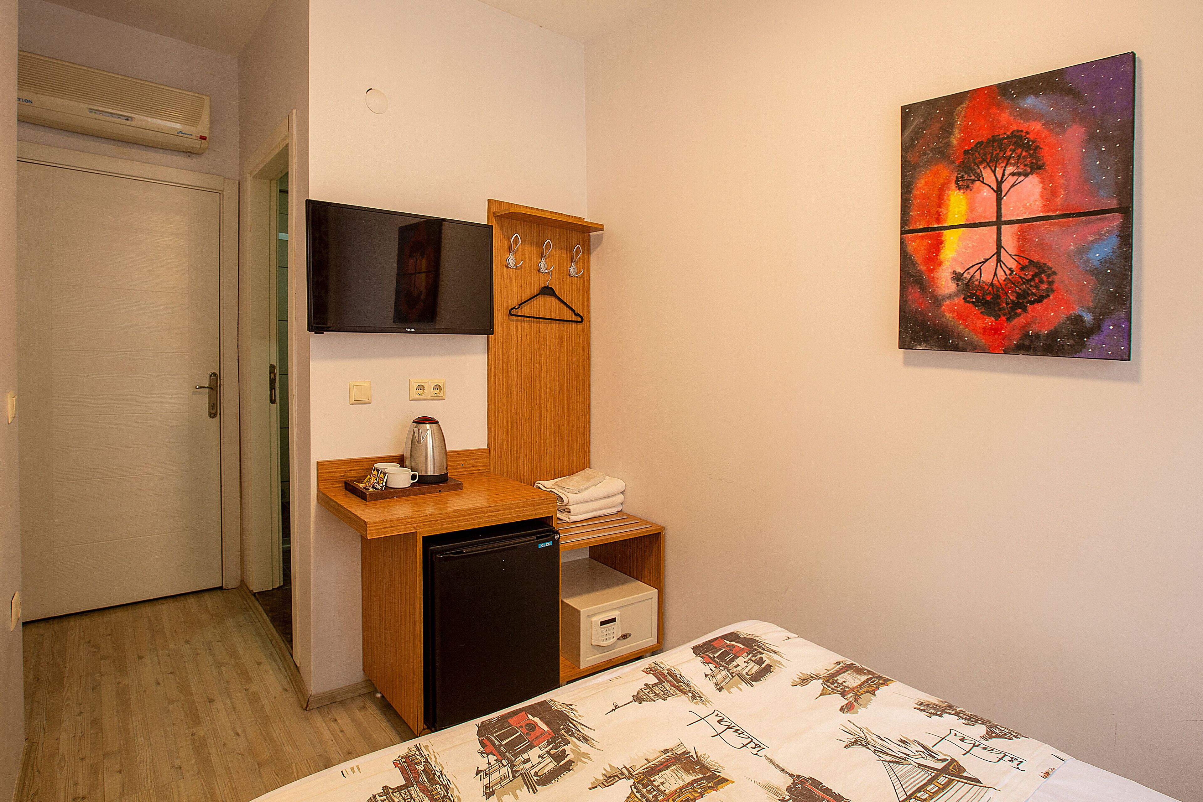 A compact hotel room with a bed, a small desk with a refrigerator, a TV, and a wall-mounted artwork.