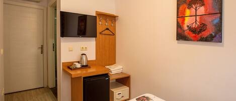A compact hotel room with a bed, a small desk with a refrigerator, a TV, and a wall-mounted artwork.