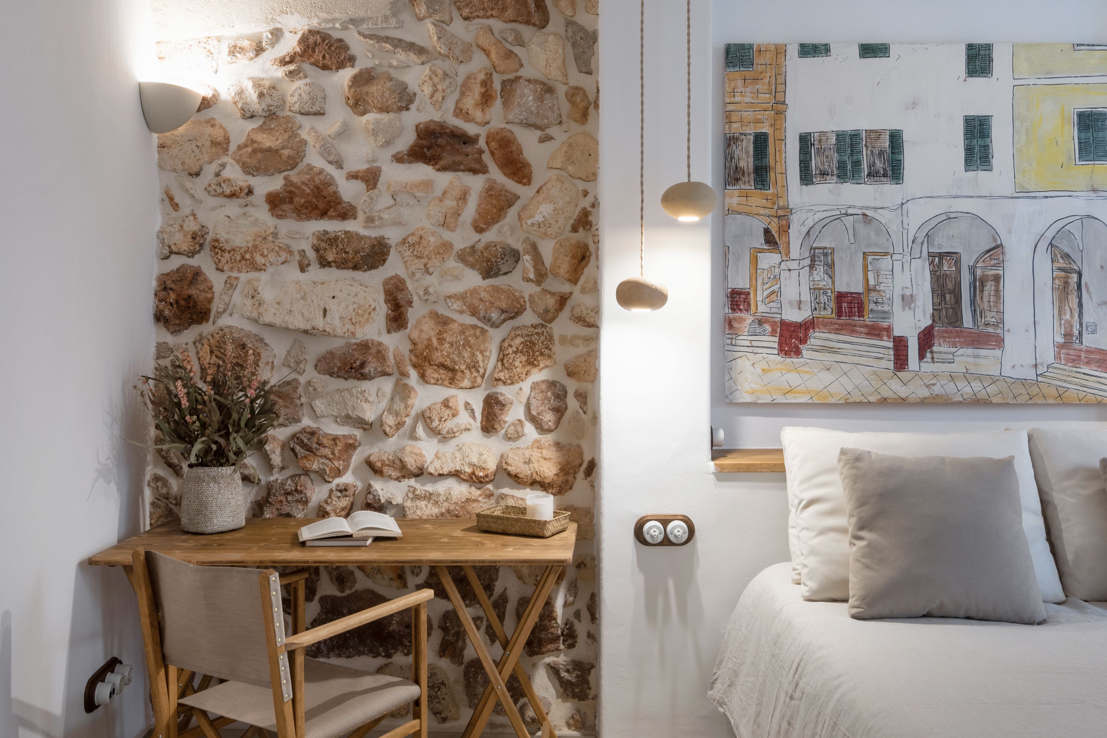 Una habitación con pared de piedra, un escritorio de madera con silla, una cama con almohadas, una lámpara de pared y un cuadro de un edificio.