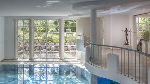 A spacious indoor swimming pool with clear glass walls, a central column, and a staircase with a railing. - ROEWERS Privathotel & Spa (Sellin)