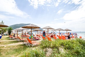 Una playa con hileras de sillas de baño naranjas y sombrillas de paja, un cielo despejado y un mar tranquilo al fondo.
