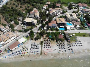Vista aérea de una zona costera con varios edificios, una playa con sombrillas y tumbonas, y una piscina.