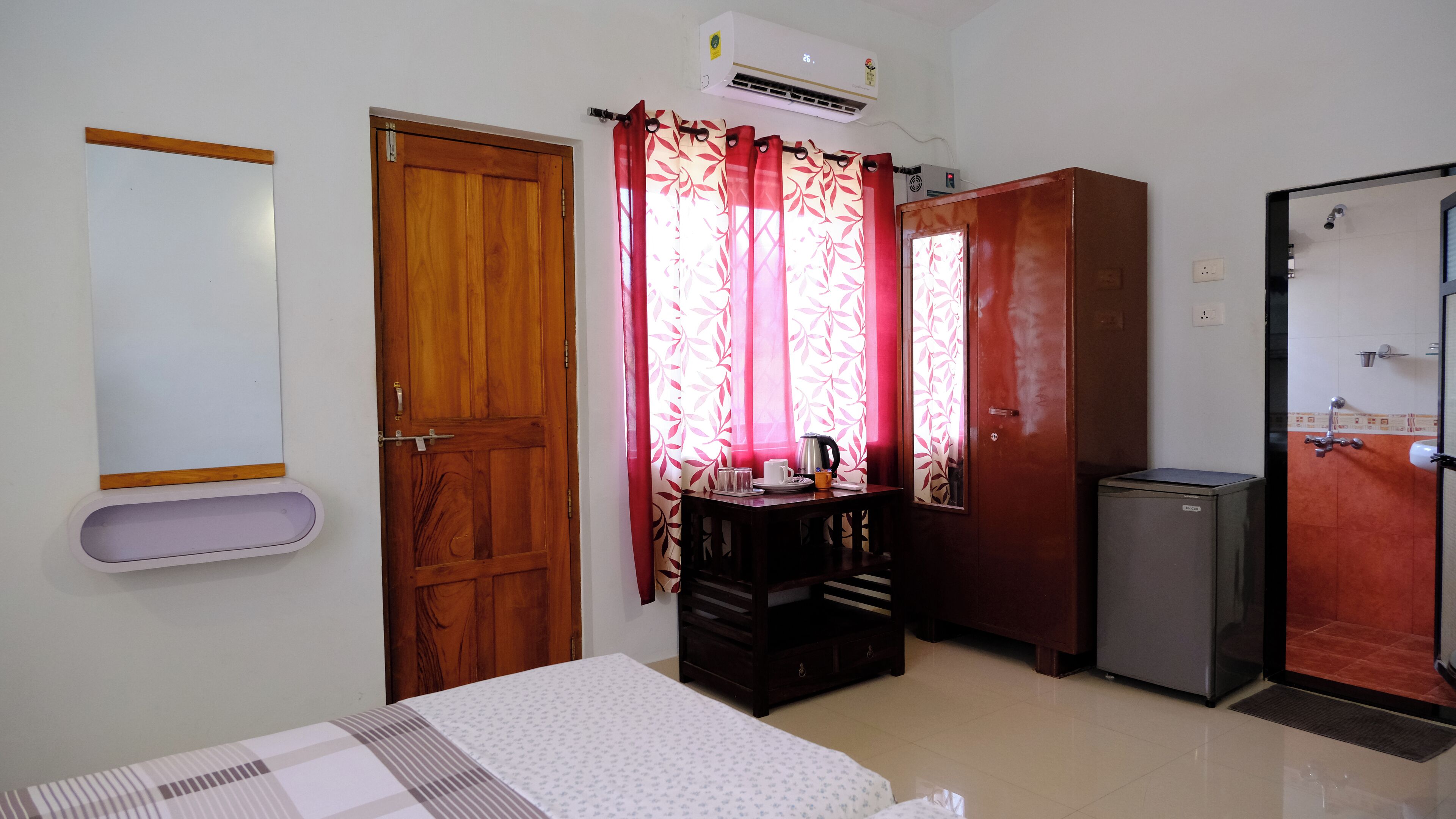 A room with a bed, a desk with a kettle, a refrigerator, and a bathroom with a shower.