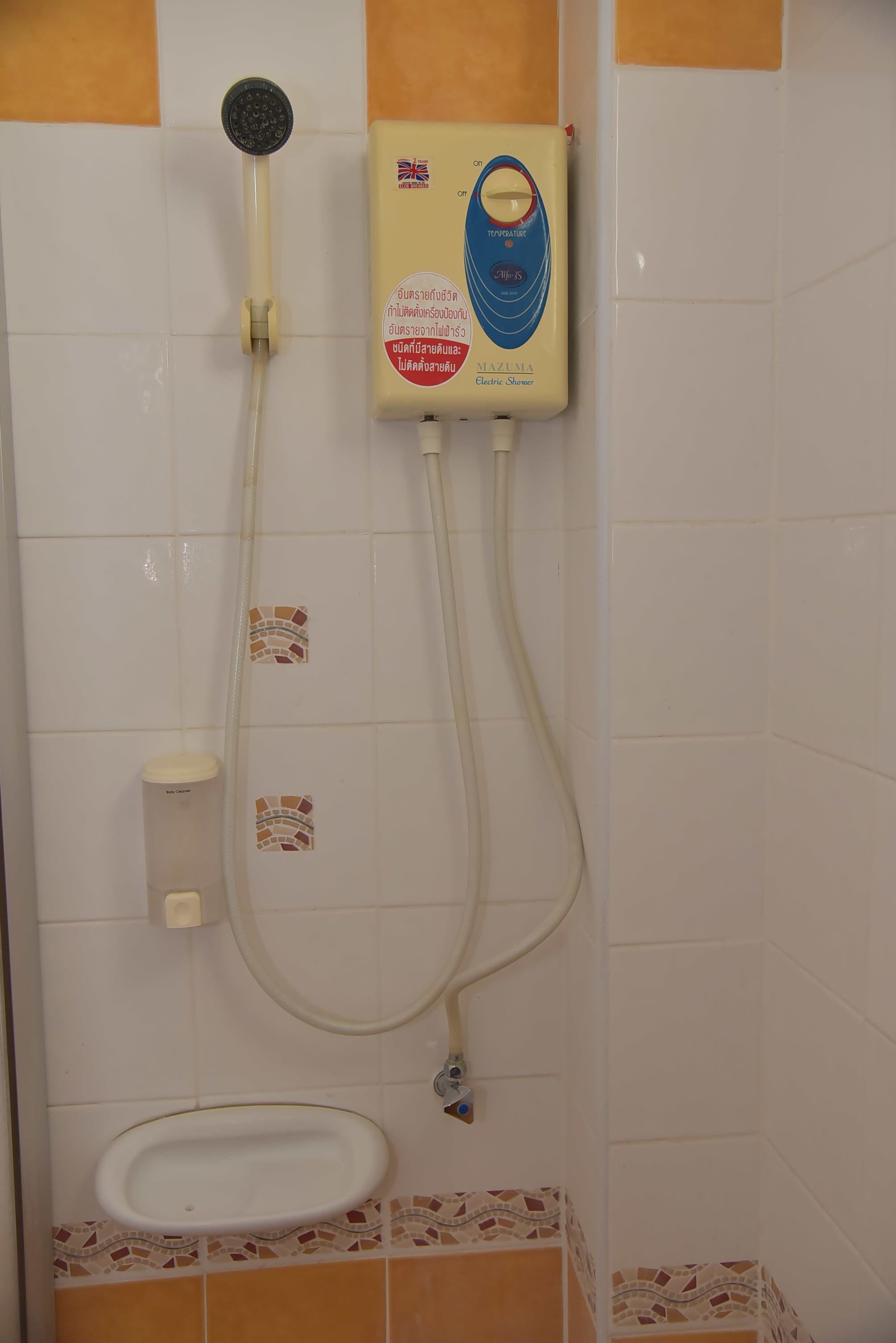A bathroom with a white tiled wall, a shower with a handheld showerhead, a wall-mounted electric shower unit, and a small sink with a mosaic tile border.