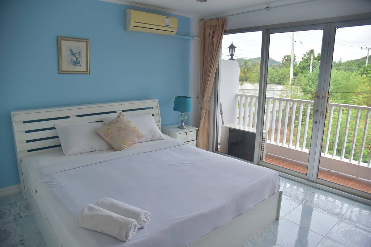 A bedroom with a bed, a nightstand, an air conditioner, a window with curtains, and a view of greenery outside.