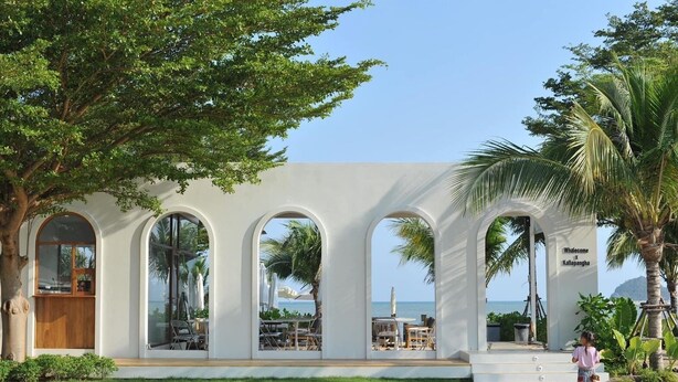 Un edificio frente a la playa con ventanas arqueadas que dan al mar, una puerta de madera y un letrero que dice "Whalecome x Kallapangha".