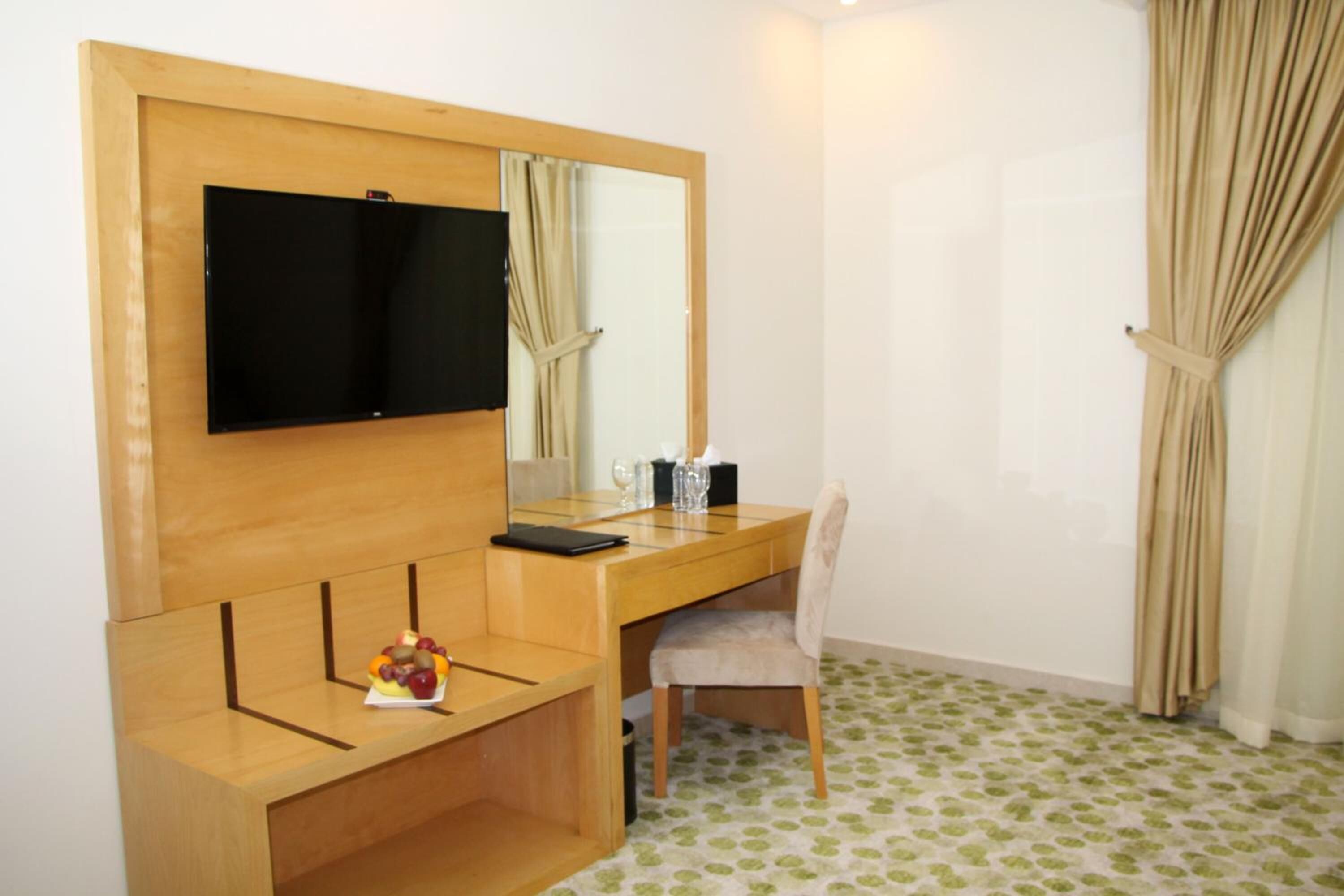 A hotel room with a wooden desk, a chair, a television mounted on the wall, a mirror, and a small fruit bowl on the desk.