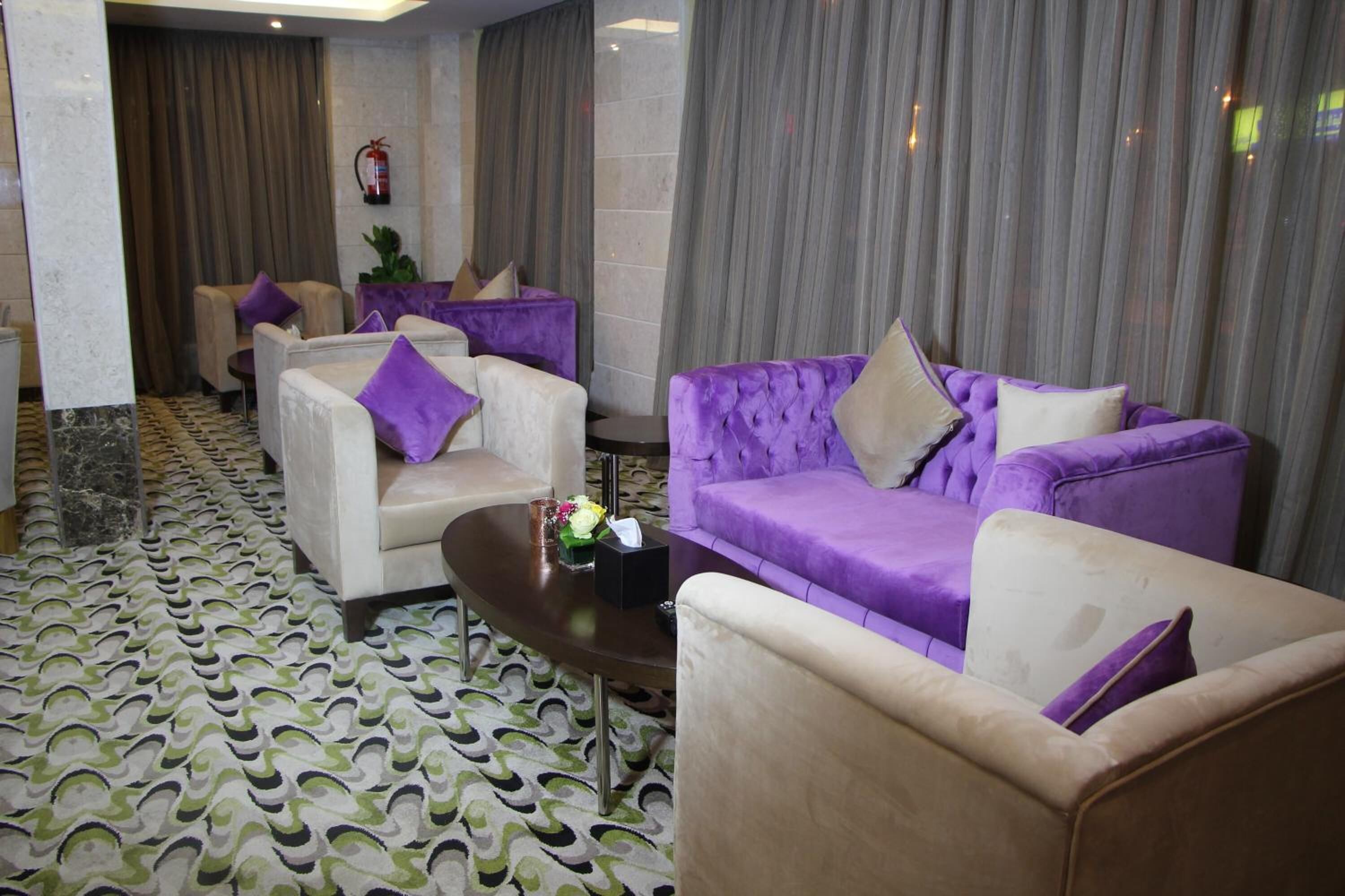 Eine moderne Lobby mit einem gemusterten Teppich, violetten und beigefarbenen Sofas, einem kleinen Tisch mit einer Vase und einem Feuerlöscher an der Wand.