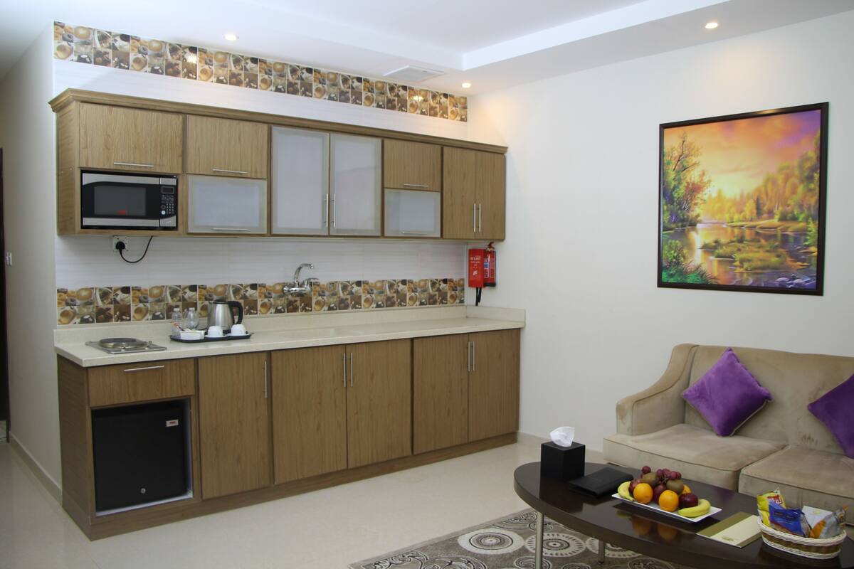 Eine kleine Küche mit Holzschränken, einer Mikrowelle und einem Spülbecken. Auf der Arbeitsplatte befinden sich ein kleiner Kühlschrank, eine Kaffeemaschine und eine Obstschale. Im Wohnbereich steht ein beiges Sofa mit violetten Kissen, und ein Landschaftsbild hängt an der Wand.