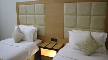 A hotel room with two beds, each with white bedding and decorative pillows. The headboard is upholstered in a light beige fabric, and there is a wooden nightstand between the beds.