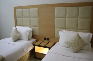 A hotel room with two beds, each with white bedding and decorative pillows. The headboard is upholstered in a light beige fabric, and there is a wooden nightstand between the beds.