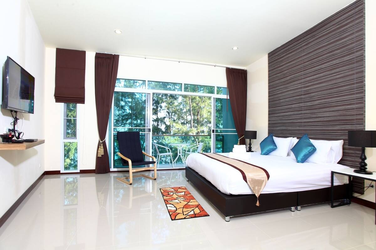 Une chambre d’hôtel moderne dotée d’un grand lit, d’une télévision, d’un balcon avec une chaise et d’une vue sur les arbres.