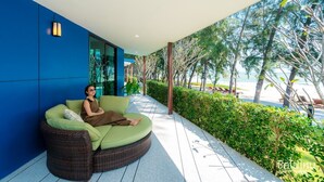A modern outdoor seating area with a curved green sofa, a person seated, and a view of the beach and trees. - Sea & Sea Villa Resort Sangaroon (Thap Sakae)