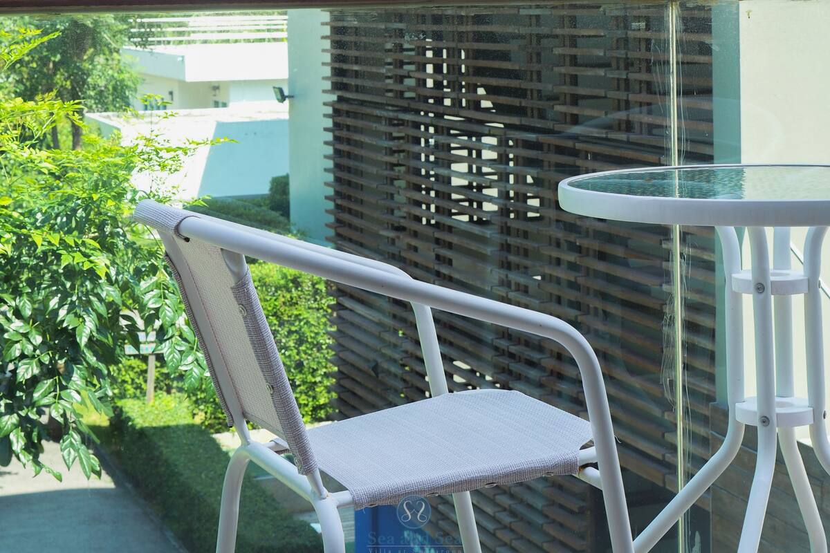 Un balcon avec une chaise et une table, donnant sur une zone verdoyante et luxuriante, avec une pancarte indiquant « Villa Sea and Sea à Sanggau ».