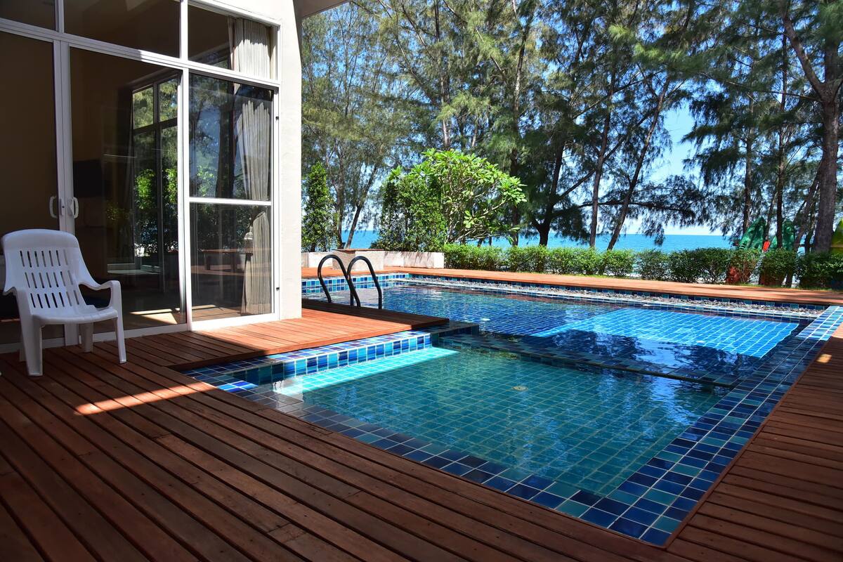 Une terrasse en bois avec une piscine, une chaise blanche et une porte vitrée donnant sur une pièce.