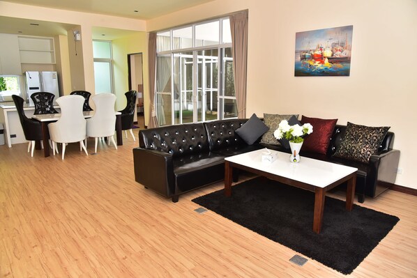 A modern living room with a black leather sofa, white dining table, and a large window with curtains.
